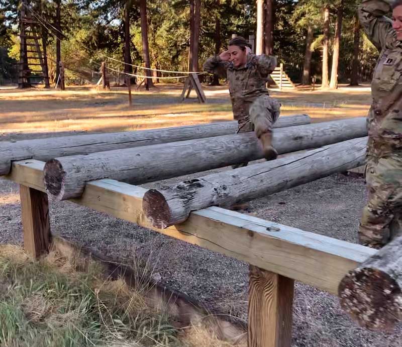 Woman in uniform maneuvers over logs as part of a drill. 