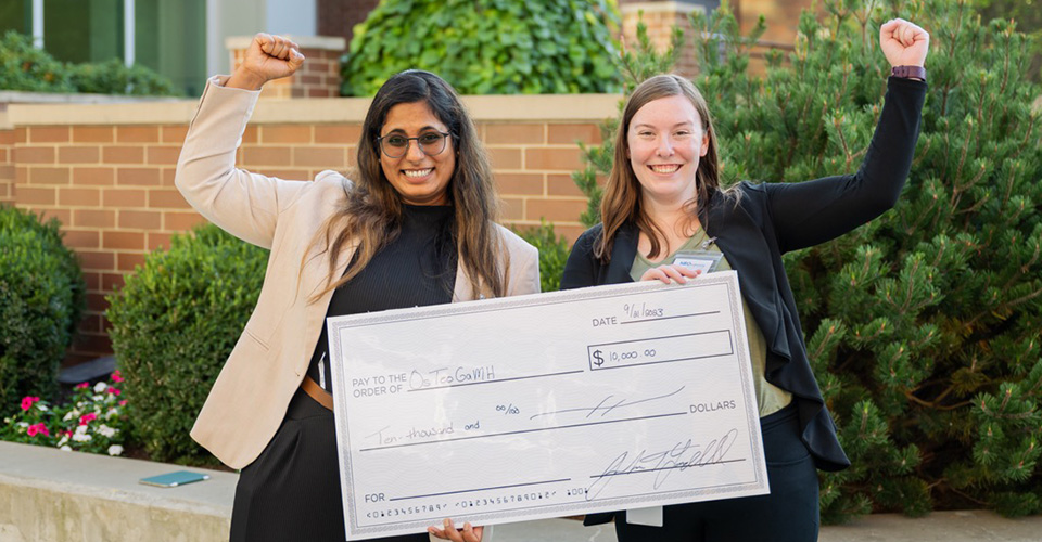Two smiling women, who won the 2023 competition hold a large check while raising their fists.