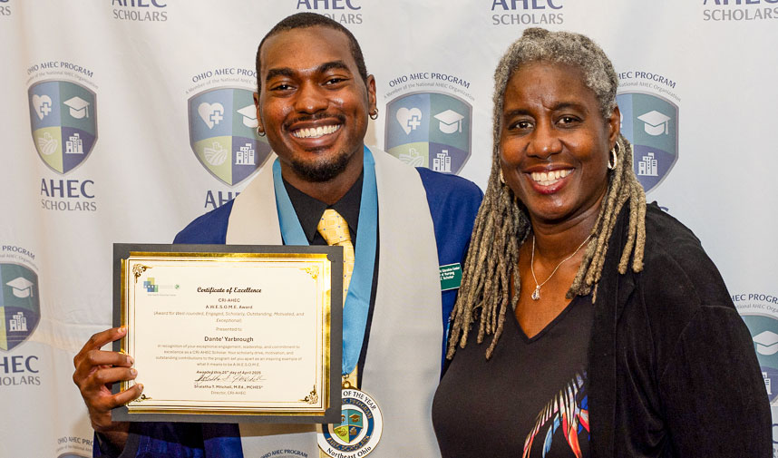 Smiling young man poses with smiling older women while holding a certificate against an AHEC backdrop