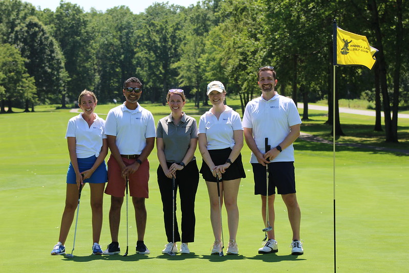 a group of five golfers poses on the green next to a hole with a yellow flag
