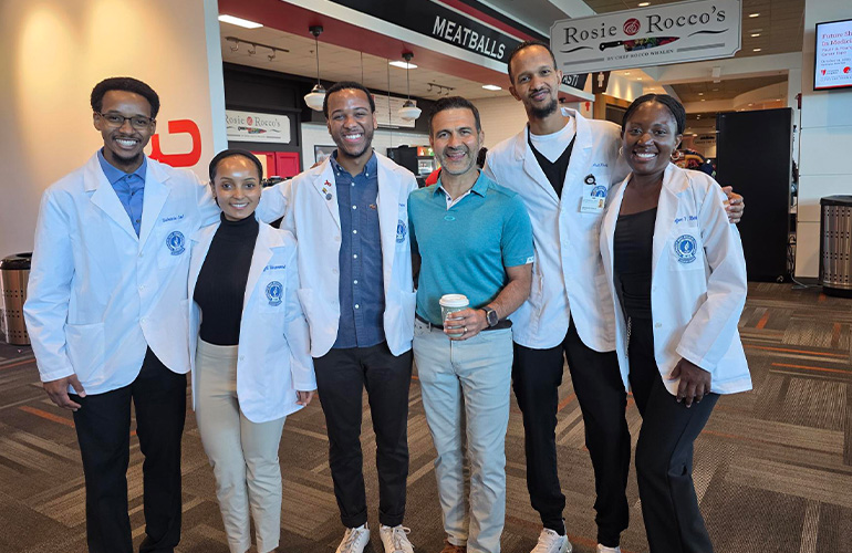 Group of young people in white coats pose with man wearing a teal polo shirt