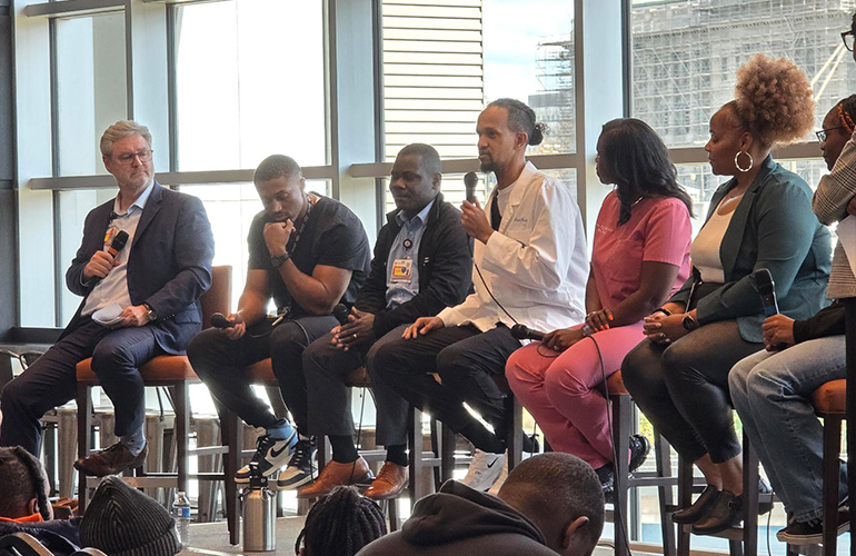 Young man wearing a white coat and holding a microphone sits in the center of a discussion panel 
