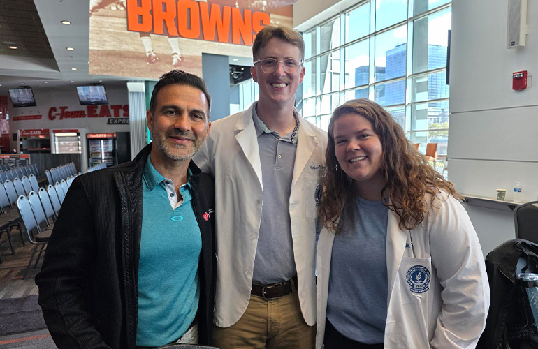 man in teal polo shirt poses with two young people in white coats