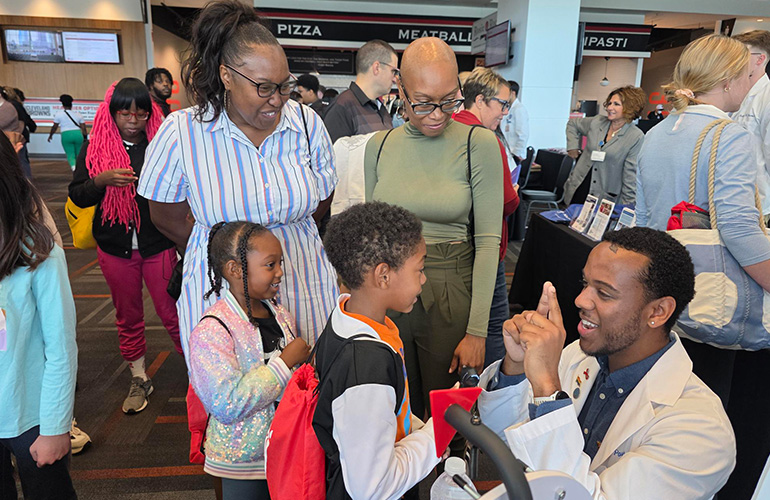 Young man in white demonstrates something to two children while their parents look on