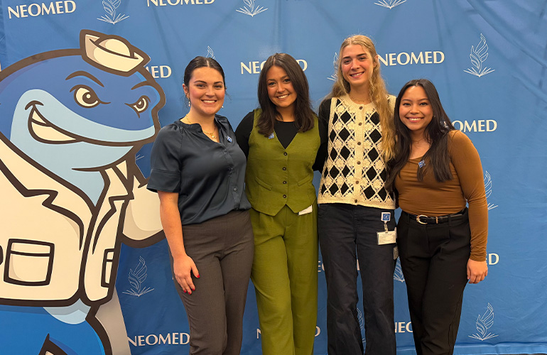 Group of four young women pose in front of NEOMED backdrop with Nate the Walking Whale