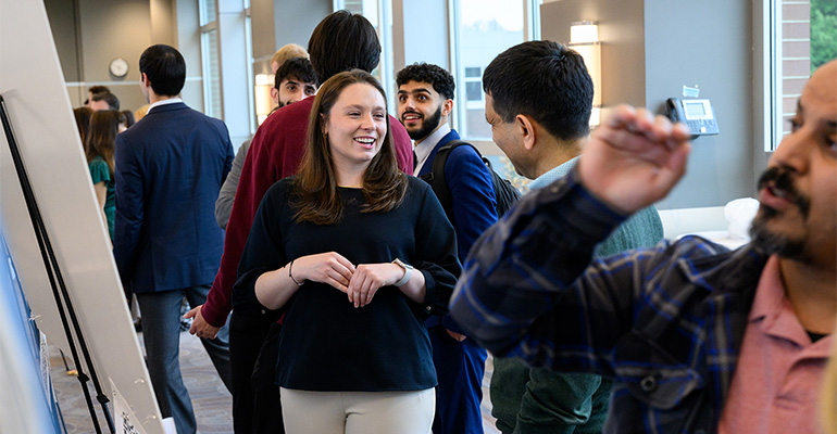Young woman in a dark sweater smiles as she explains her scientific poster to a visitor
