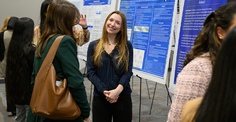 Young woman in a dark sweater smiles in front of an academic poster