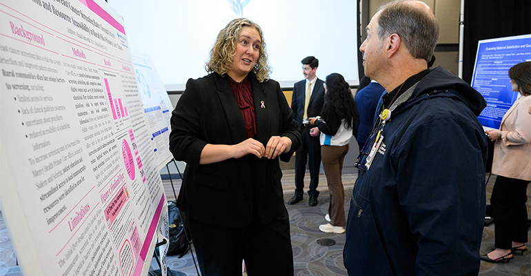 Blond woman in a black suit explains her scientific poster to a man in a blue jacket