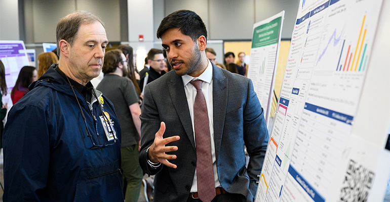 Young man in a suit and tie explains his scientific poster to a visitor