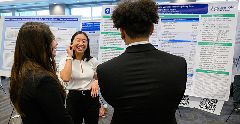 young woman in cream-colored top smiles while explaining her scientific poster to two onlookers