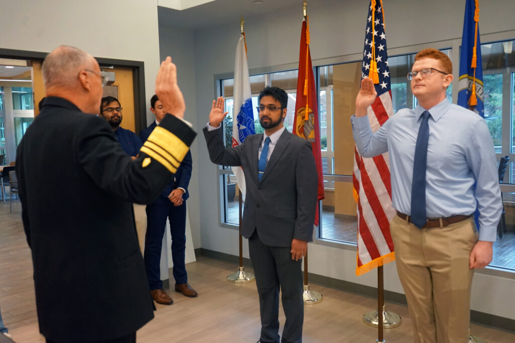 Two young men with their right hands raised are sworn into their commission by a man in a Navy uniform