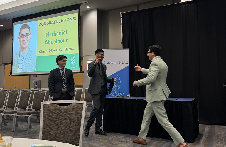 Man in suit walks toward podium to receive a certificate. Screen in the background shows a slide of Nathaniel Abdelnour