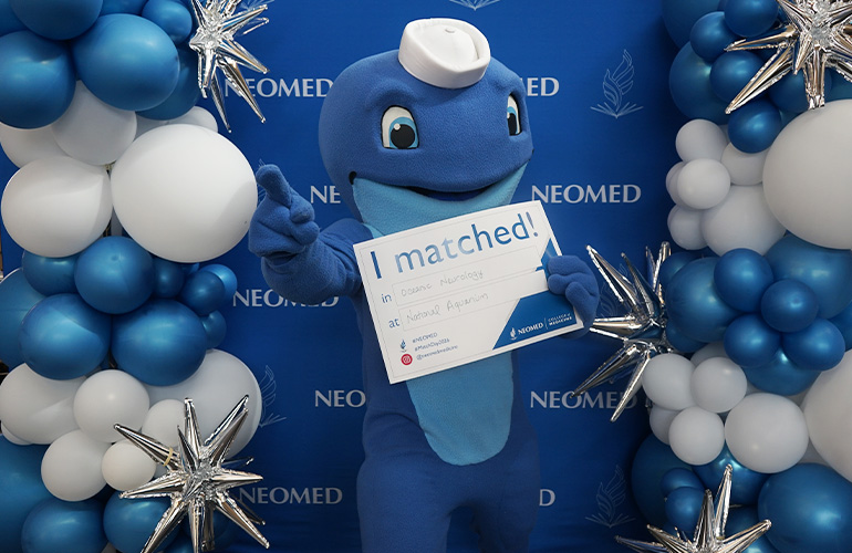 Nate the Walking Whale holds a sign saying "I Matched" in front of a NEOMED backdrop. The sign says he matched in oceanic neurology at the National Aquarium.