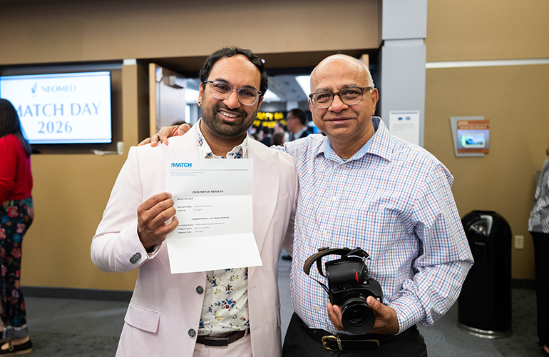 A young man holds up a letter showing that he matched into residency. An older man poses next to him with his hand resting on the young man's shoulder.