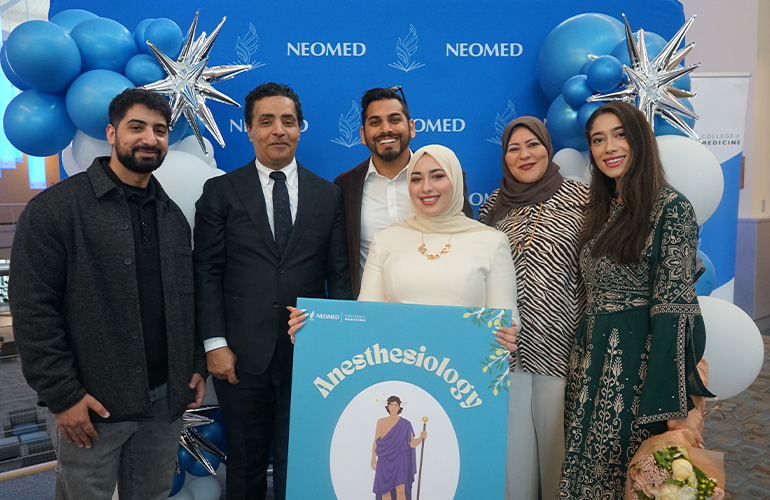 A young woman holding a poster depicting anesthesiology is surrounded her family in front of a NEOMED backdrop.