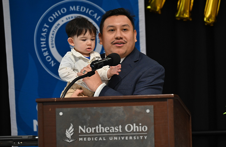 A man stands at a NEOMED podium holding a baby boy.