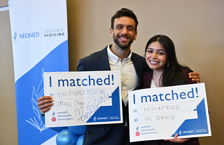 A man and a woman hold signs saying "I Matched" while standing in front a NEOMED College of Medicine banner. He matched emergency medicine at U Mass and she matched pediatrics at UC Davis