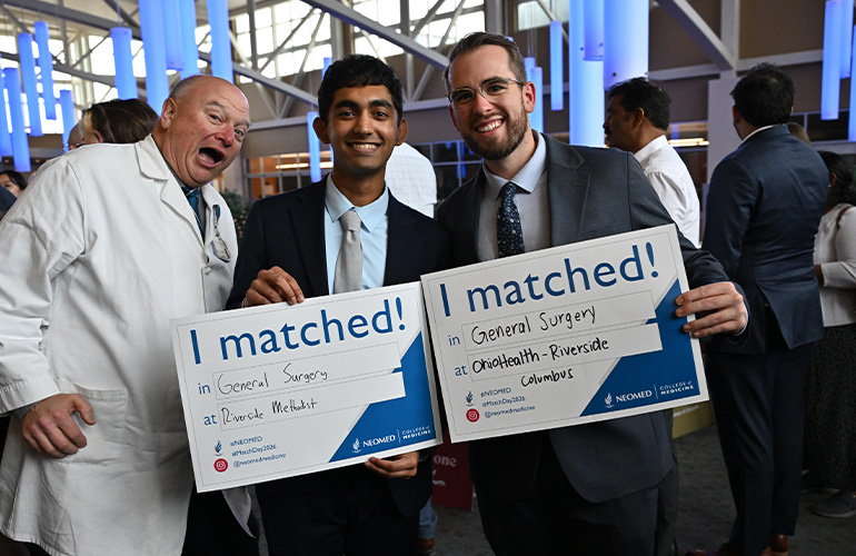 Two young men hold signs saying "I matched" while a man in a white coat makes a silly face behind them