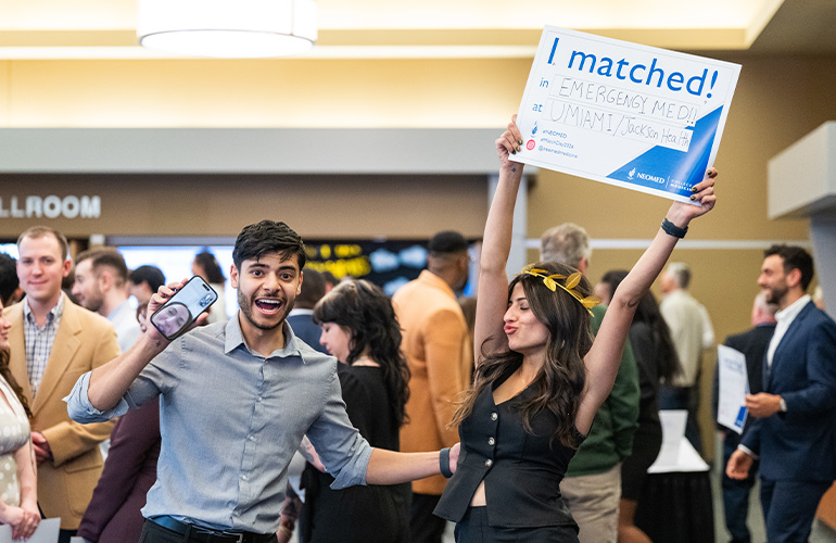An excited young man holds up a cell phone camera while standing with a proud young woman holding a sign above her head that says "I matched"