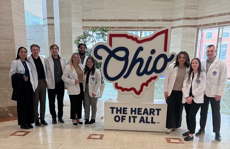 Group of pharmacy students in white coats pose with a large sign reading "Ohio, the heart of it all"