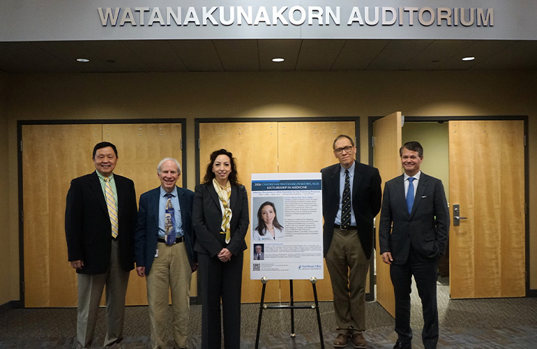 A group of five people in business attire pose beneath a sign reading "Watanakunakorn Auditorium"