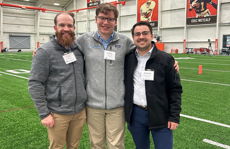 Three men in business casual pose on a training field
