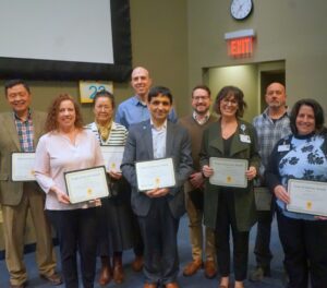 Group of people in business casual attire pose with certificates of award