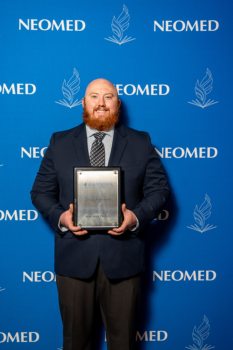 Jacob Sweet, in suit and tie, poses with an award plaque in front of a blue NEOMED backdrop