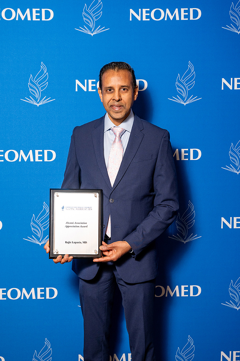 Rajiv Lapasia, in suit and tie, poses with an award plaque in front of a blue NEOMED backdrop