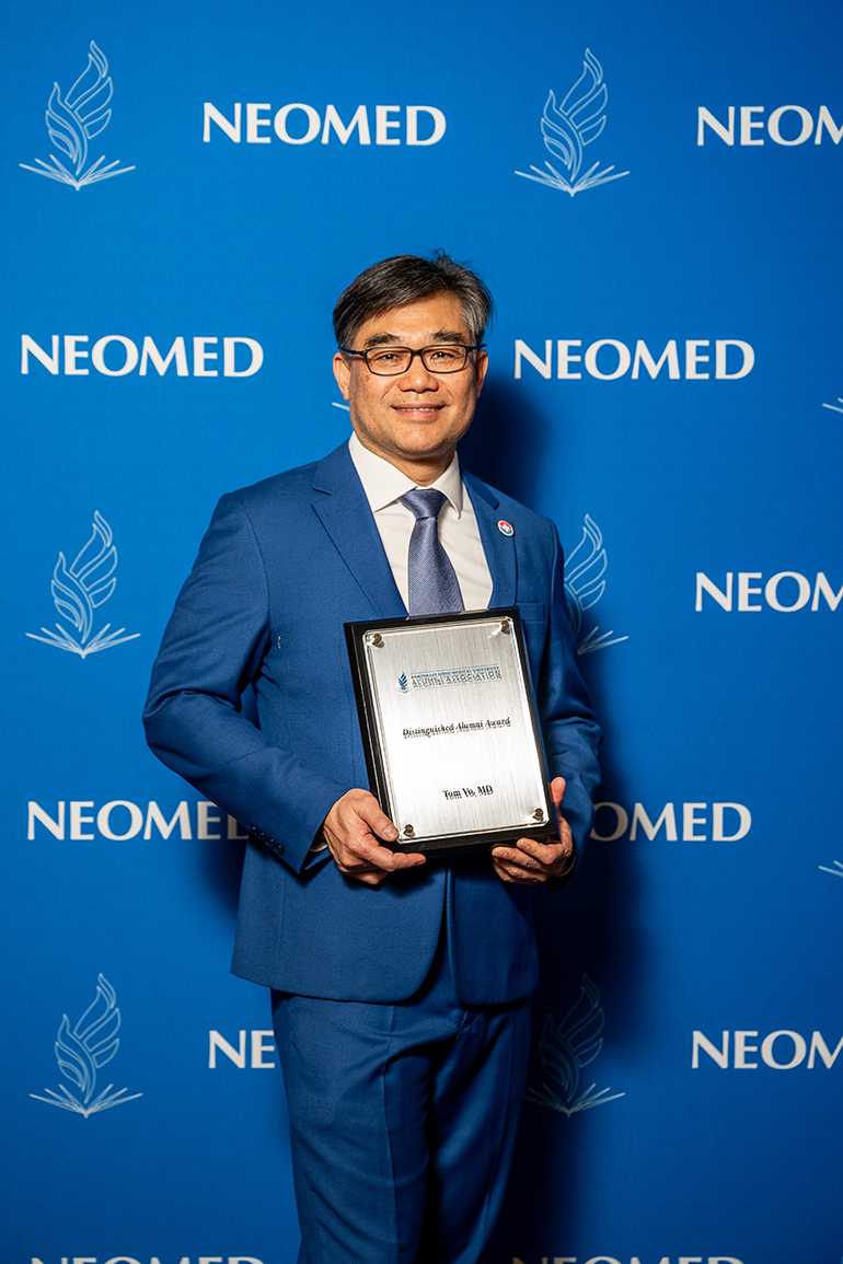 Tom Vo, in suit and tie, poses with an award plaque in front of a blue NEOMED backdrop