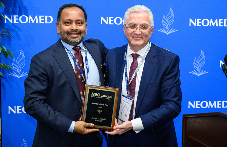 Two men in suits pose with a plaque for "mentor of the year" in front of a blue NEOMED backdrop