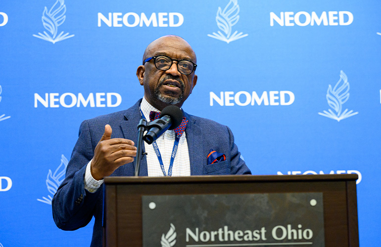 Dr. Michael Forbes speaks at a podium in front of a blue NEOMED background