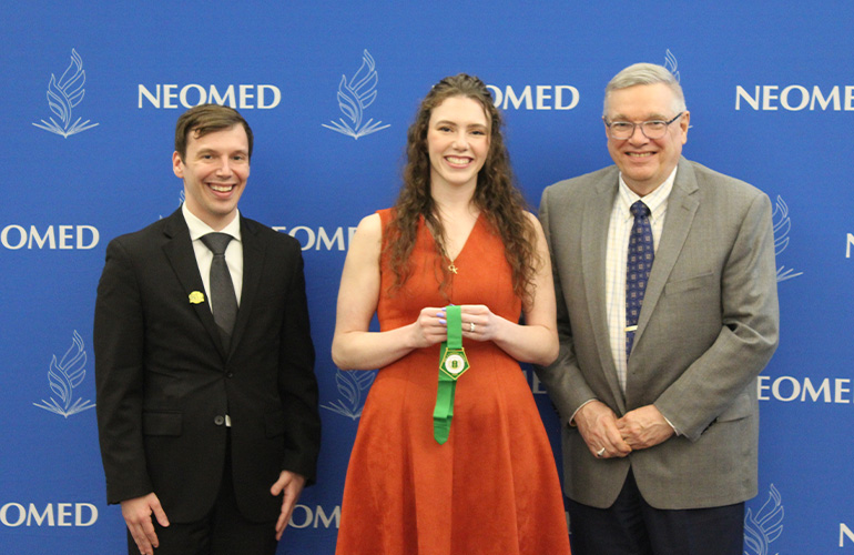 Two men in suit coats pose on either side of a young woman wearing a dress and holding a green ribbon. They pose in front of a blue NEOMED backdrop.