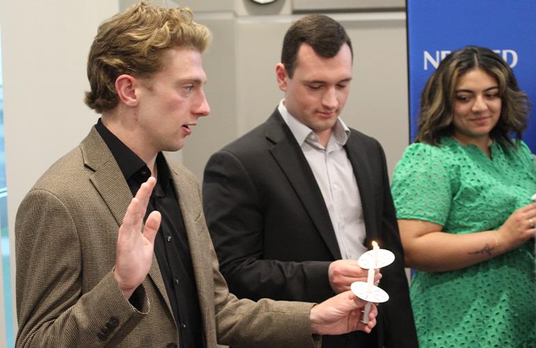 A young man holds up his hand as though taking an oath, while another young lights a candle that he is holding. A young women looks on.