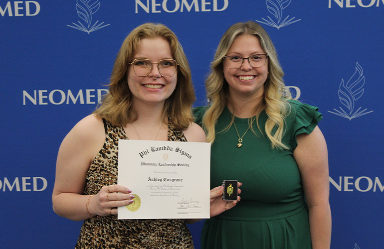Two women wearing glasses and business attire pose with a certificate in front of a blue NEOMED backdrop