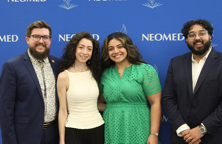Four young adults in business casual attire pose in front of a blue NEOMED backdrop