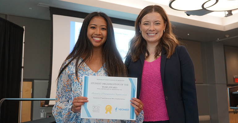 Two women smile at the camera. The woman on the left holds an awards certificate