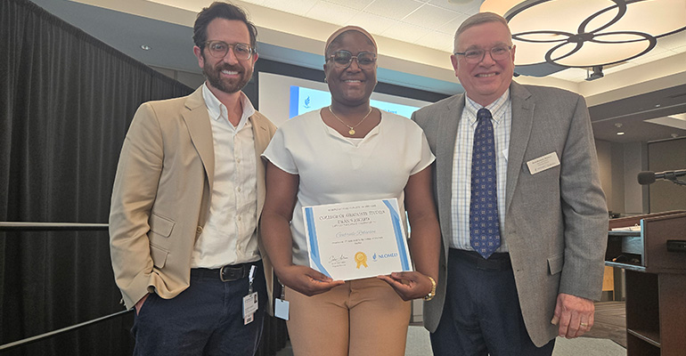 Two men and a woman smile at the camera. The women, center, holds an awards certificate