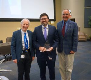 Three men in suits pose in a large conference hall in front of a screen