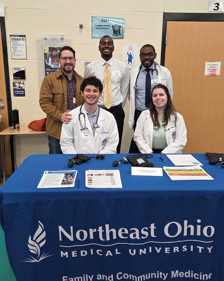 Two young people in white coats are seated at a table with a Northeast Ohio Medical University - Family and Community Medicine tablecloth. Three men are standing behind as they pose for the photo