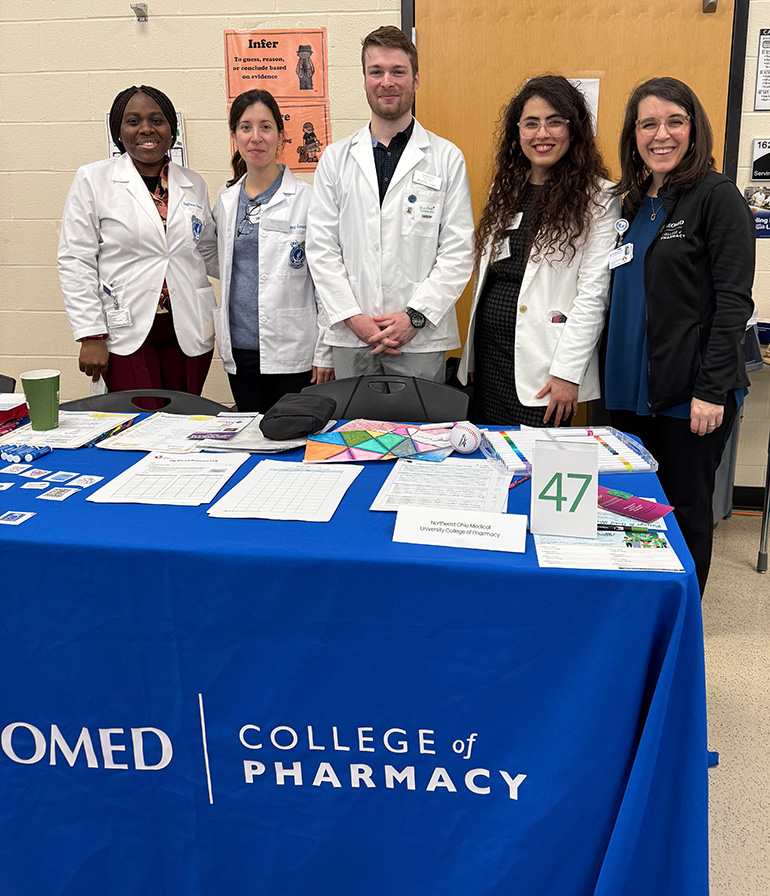 Five people in white coats or other NEOMED-branded apparel stand behind a table fdraped by a NEOMED College of Pharmacy tablecloth