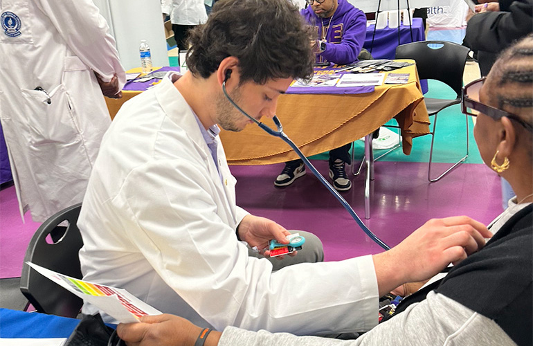 A young man in white coat checks the blood pressure of a woman wearing glasses 
