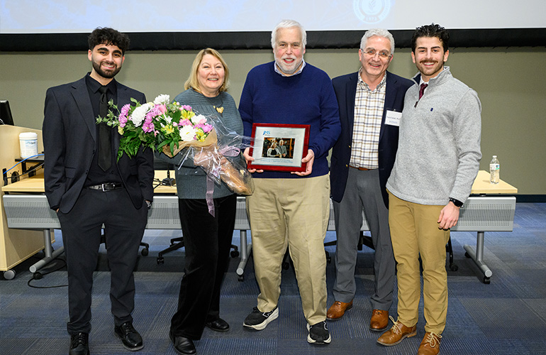 a woman and man in business casual attire pose with three men while holding a bouquet of flowers and a plaque