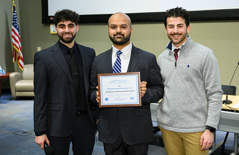 Three young men in business attire pose in a large meeting space. The man in the center holds a recognition plaque