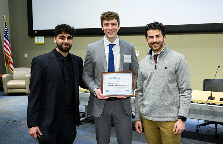 Three young men pose in a large meeting room. The man in the center holds a recognition plaque