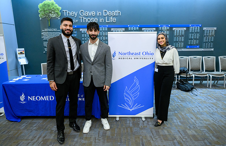 Two men and a woman in business attire stand on either side of a NEOMED pop-up banner in front of a table with a NEOMED tablecloth.