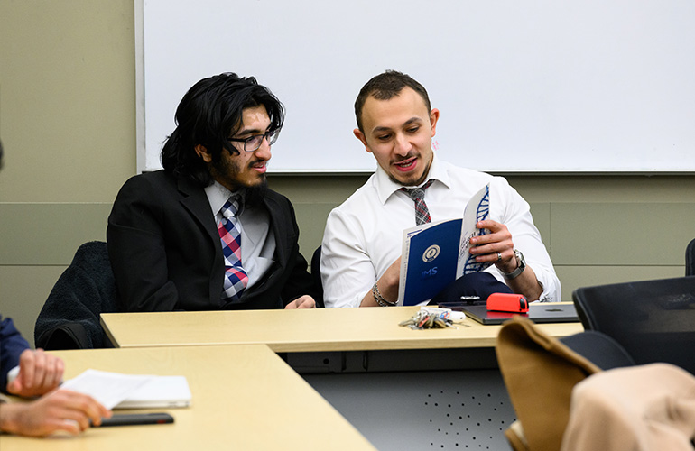Two men in business attire look at something in a program booklet while seated at a table