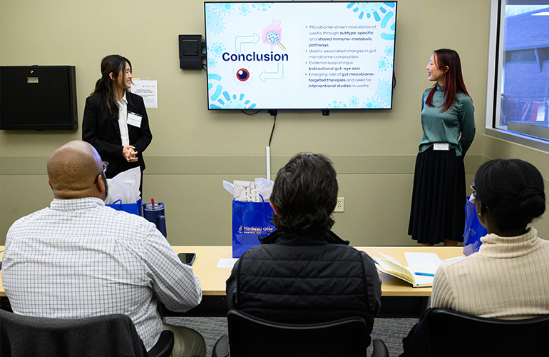 Two young women stand on either side of a conference screen, with a group of people looking on from seats