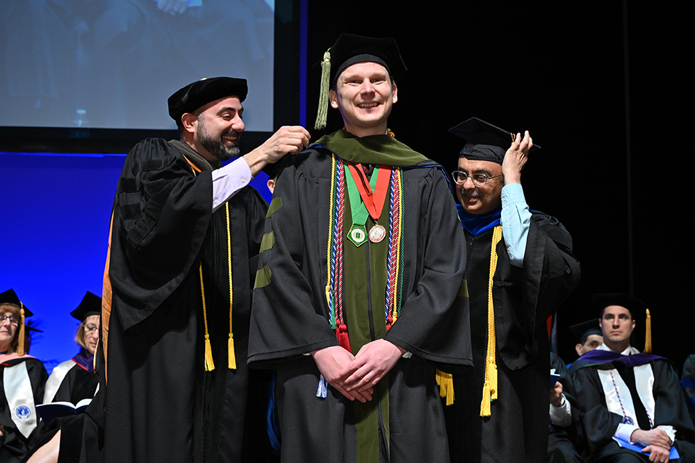 Two faculty members place a doctoral hood on a pharmacy graduate during a commencement ceremony.