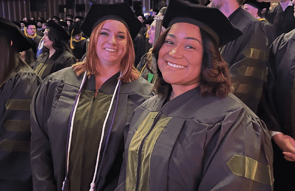Two women graduates dressed in  academic regalia smile at the camera